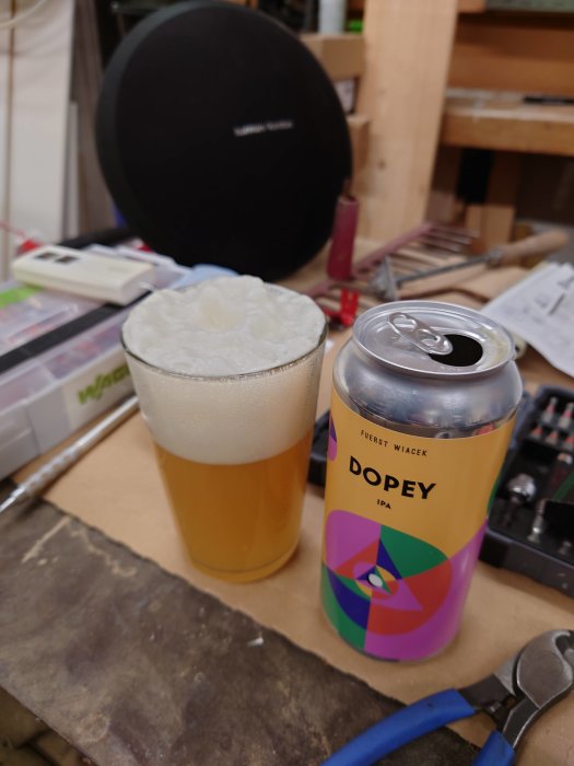 Ett ölglas med skum vid sidan av en Fuerst Wiacek Dopey IPA-burk på ett verktygsbord.