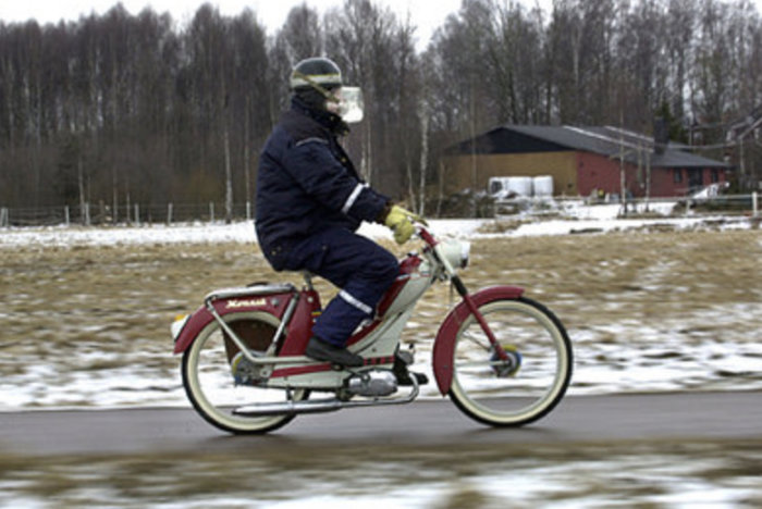 Person kör röd moped på slaskig väg med snötäckta fält och hus i bakgrunden.