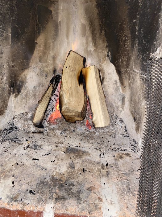 Brinnande vedträn i en eldstad med aska och sot på väggarna.