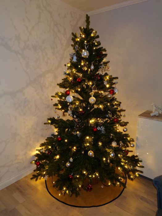 Dekorerad julgran med vita, röda och silverprydnader och tända ljus, inomhus mot en ljus vägg.