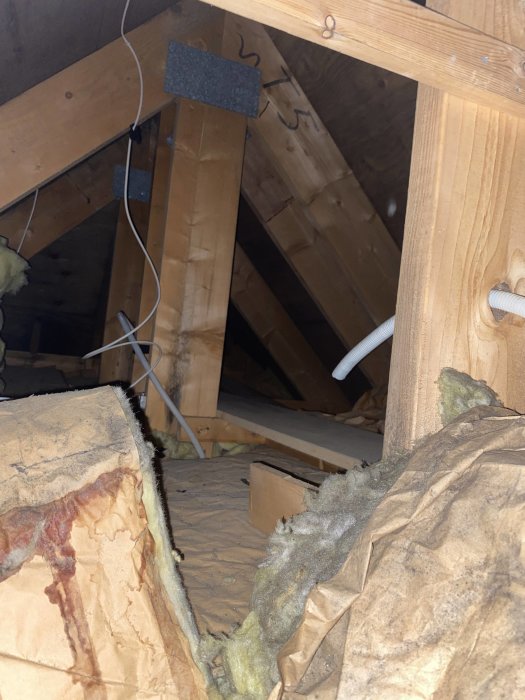 En otydlig vindsinteriör med isolering, träbjälkar och synliga kablar i svagt ljus.