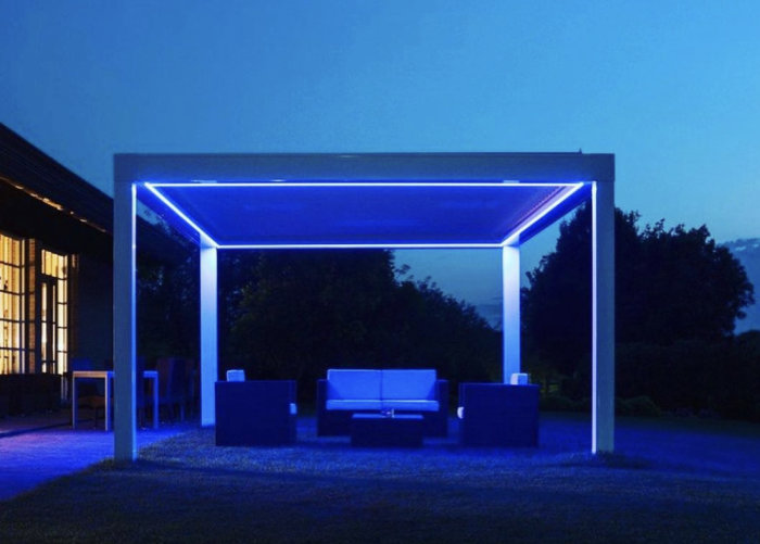 Modern pergola med blått LED-ljus över utemöbel i en trädgård vid skymning.