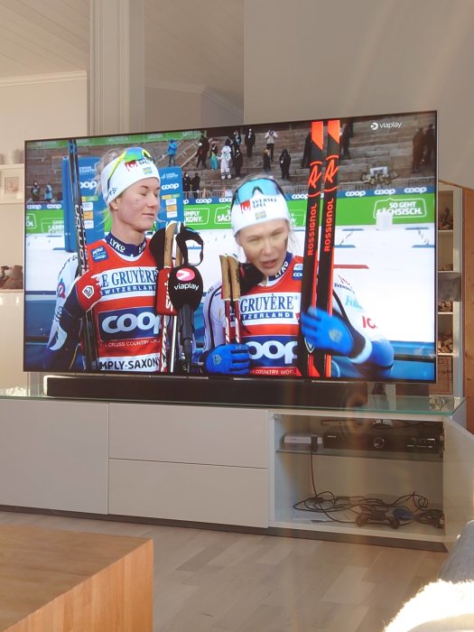 Två skidåkare i tävlingsdräkter intervjuas på en TV-skärm i ett vardagsrum.