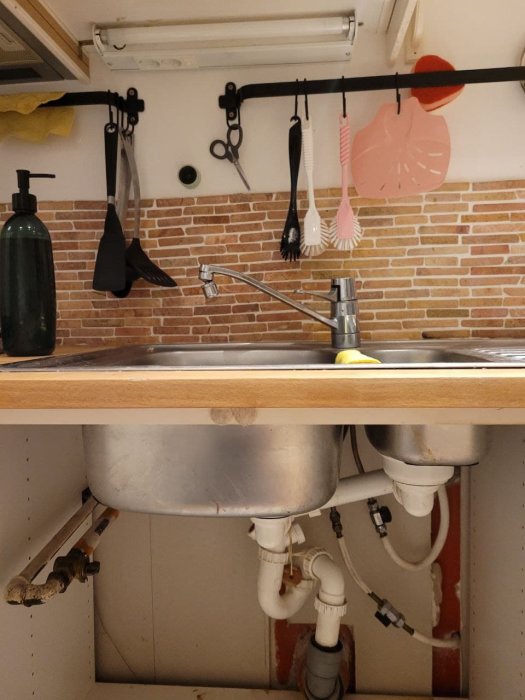 Köksskåp under diskho med synliga rör och vattenkopplingar, vägg med tegelbacksplash.