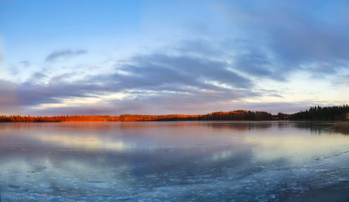 Isfrusen sjö vid solnedgång med skog i bakgrunden och reflektioner på isen.