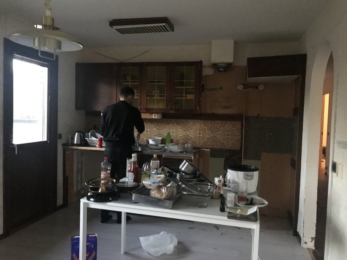 Ett kök före renovering med äldre inredning och en person som står framför fönstret.