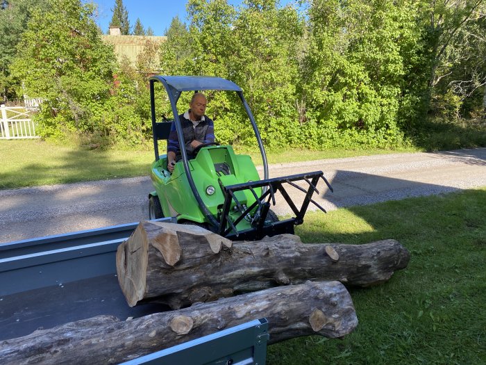 Man lastar stora trästockar på en släpkärra med hjälp av en liten grön traktor.