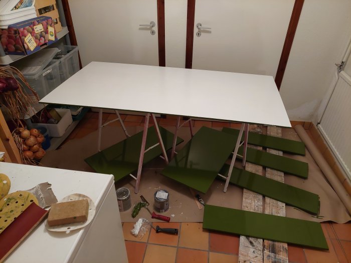 Vitmålad bordsskiva på bockar med grönmålade bordben och målartillbehör på golvet.