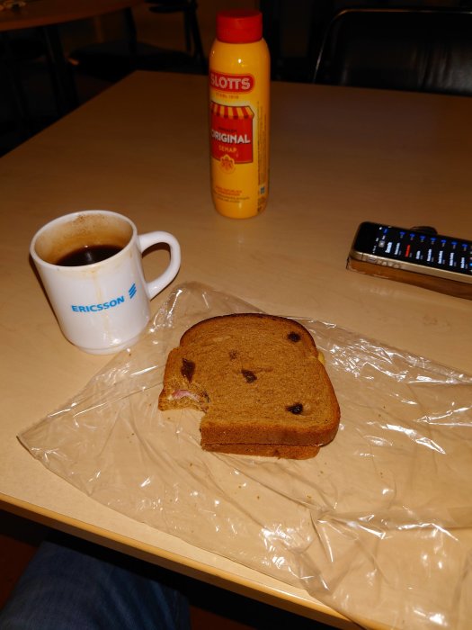 Kaffekopp, rostat bröd med russin, senapsflaska och en mobiltelefon på ett bord.