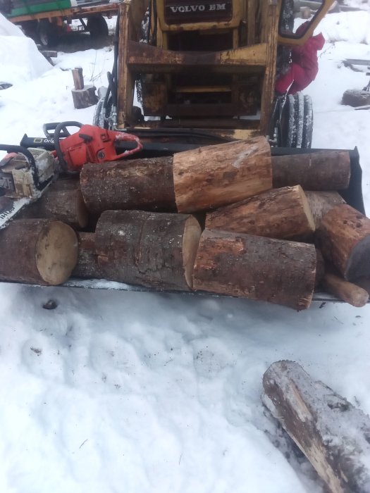 Granved i olika längder staplade på snö framför en motorsåg och en gul hjullastare i bakgrunden.