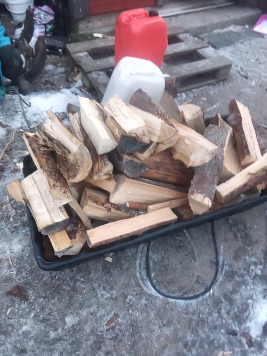 Hög med nyskuren ved på en släde framför en röd kanister, utomhus med snöfläckar på marken.