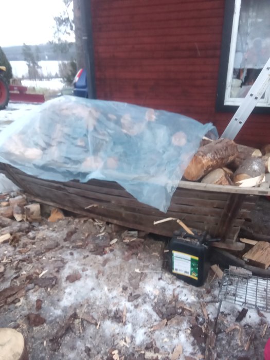 Övertäckt vedhög bredvid en stege och ett rött trähus med delvis synlig släpvagn.