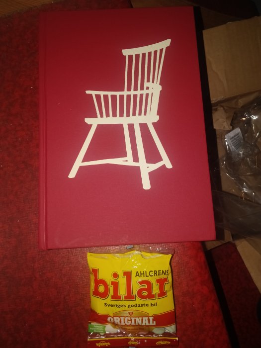 Röd bok med en vit stol på omslaget ligger ovanpå ett paket Ahlgrens bilar på ett rött underlag.