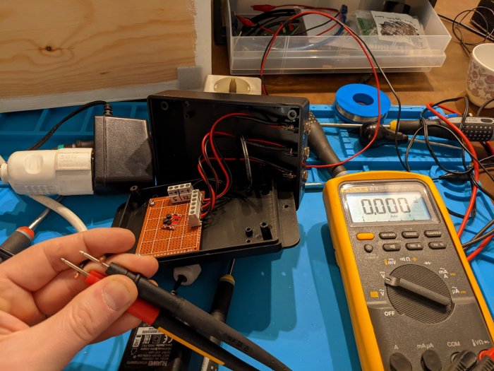 Hand håller testkablar från en multimeter med en öppen svart låda som visar elektronikkomponenter och lödkort, med verktyg runtomkring.