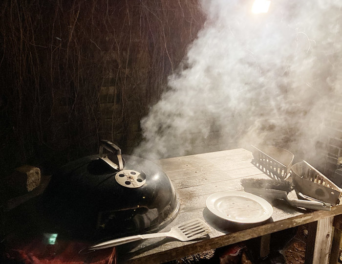 En tänd kolgrill med rykande kol på en träbänk vid skymning, med grillverktyg och en tallrik.