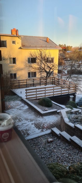 Utsikt mot ett gult hus och trädgård med lätt snötäcke i gryningen eller skymningen.