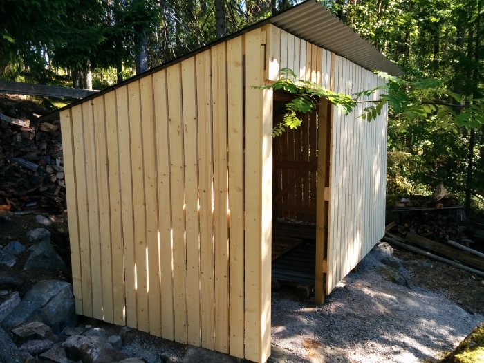 Nybyggt vedförråd med öppen sektion för ventilation, omringat av träd och staplad ved.