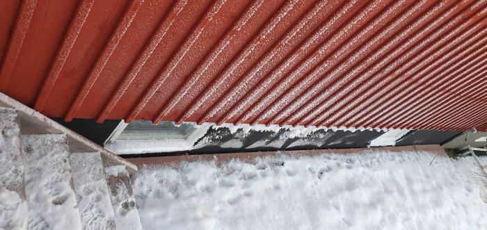 Röd takplåt med snö, slangen går in under trappen vid husgrund mot snötäckt mark.