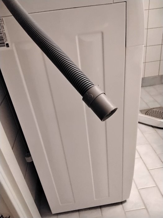 Tvättmaskinsslang hängande lös ovanför ett vitt golv med kakelvägg i bakgrunden, fråga om montering.