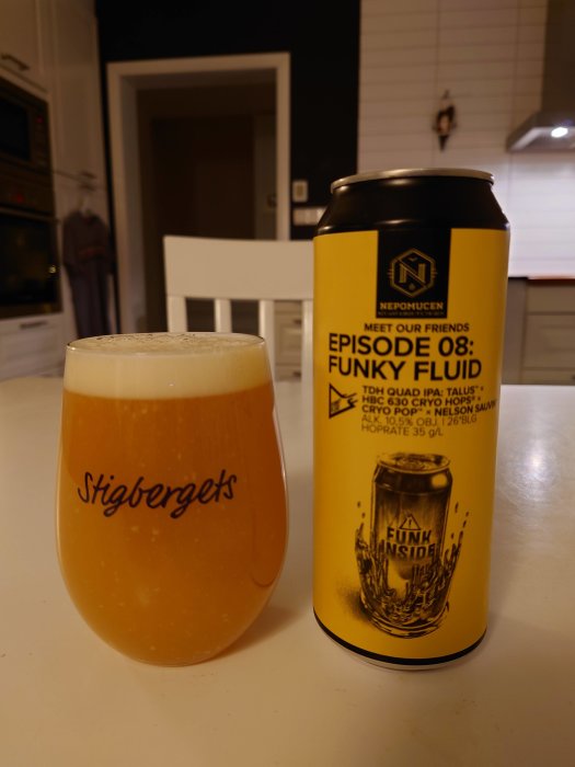 Gul ölburk bredvid ett glas med öl i ett kök, 'FUNKY FLUID' på burken, heminredning i bakgrunden.