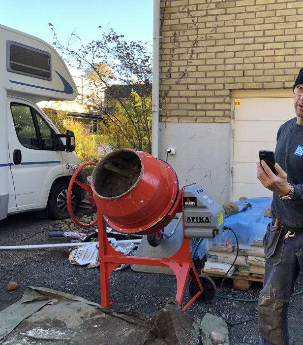 Person som använder en mobiltelefon bredvid en röd betongblandare utomhus vid en byggarbetsplats.