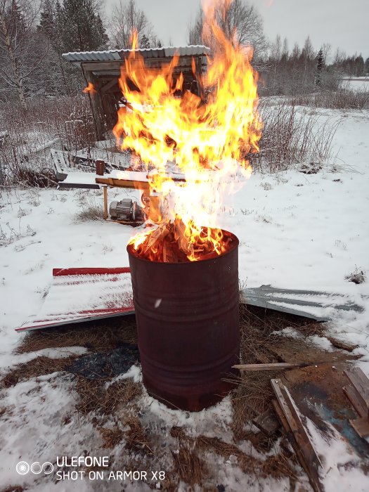 En alt-text kan lyda: Brinnande eld i en oljetunna utomhus med snö och byggmaterial runt omkring.