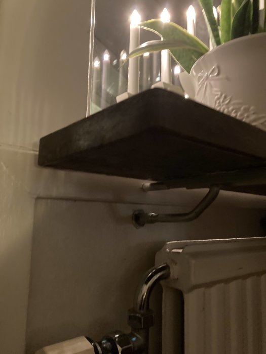 Svart hyllkonsol fäst under trähylla ovanför vit element med värmeljus och växt på hyllan.