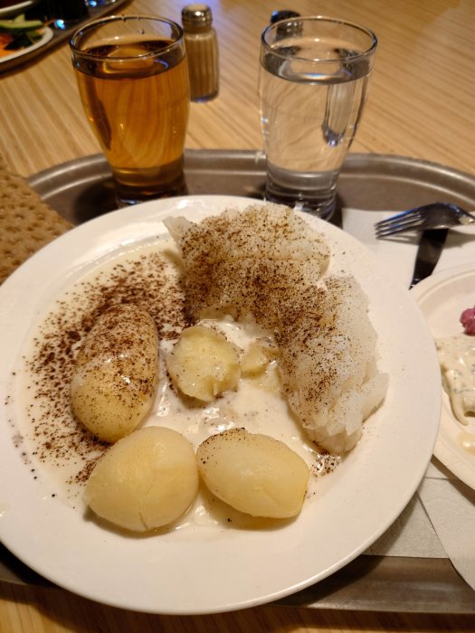 Tallrik med lutfisk toppad med vitpeppar, potatis, smält smör och serveras med öl och vatten.