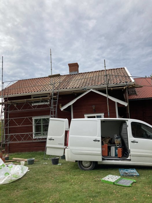 Träställning på taket vid en röd stuga för renovering av låg skorsten, verktyg och byggmaterial framför en vit skåpbil.