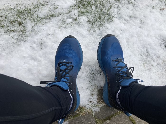 Person i svarta byxor och blåa vandringskängor står på snötäckt mark.