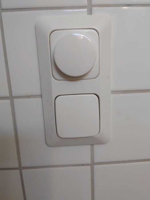 Väggmonterad strömbrytare med vit knapp och plastlock, installerad på kaklad vägg.