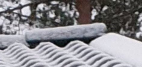 En yttersta nockpanna täckt med snö på ett tak med vågformade takpannor, träd i bakgrunden.