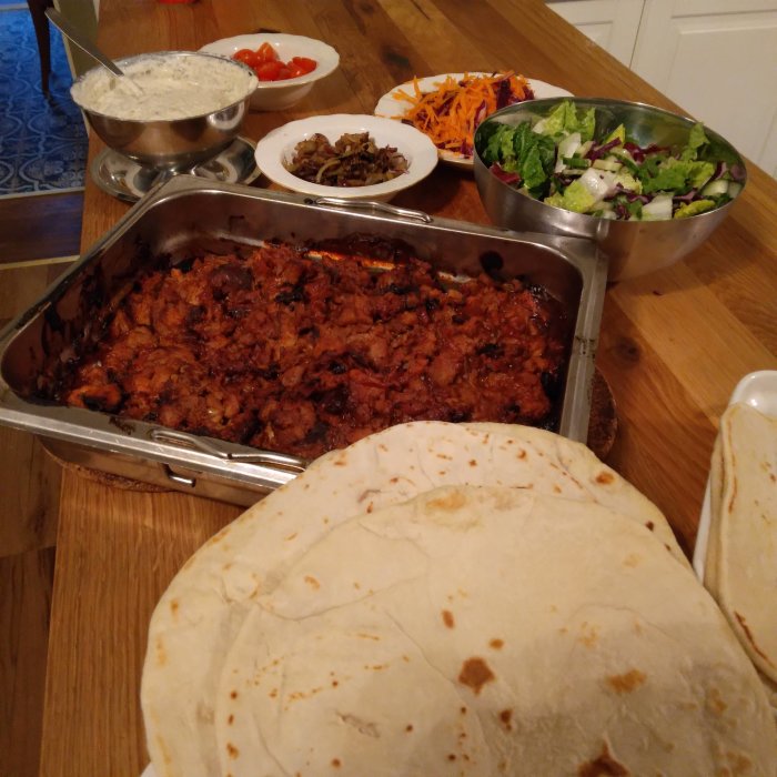 Hemgjord pulled pork i form, tacobröd och sallader på ett matbord.