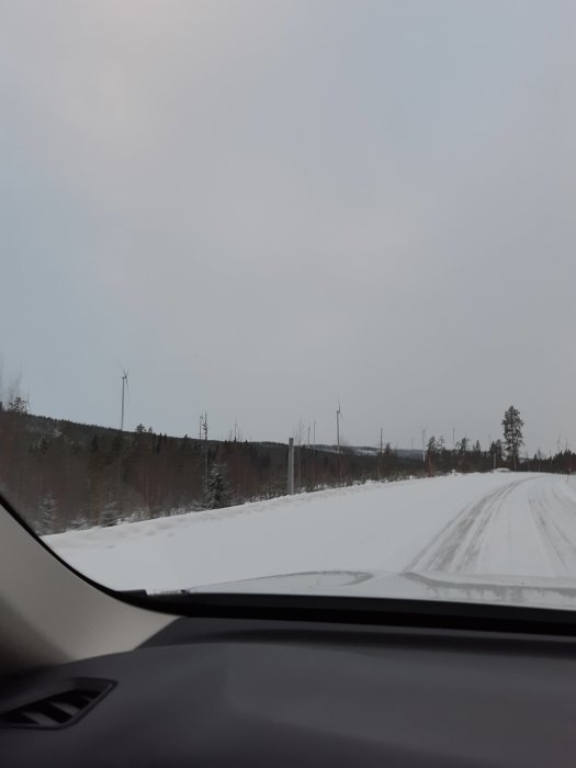 Vinterväg i norrländsk landskap med vindkraftverk i horisonten, sett från bilens passagerarsida.