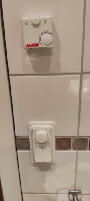 Badrumsvägg med två strömbrytare, en för golvvärme med reglage och en dimmer för taklampor, samt en mosaiklist.
