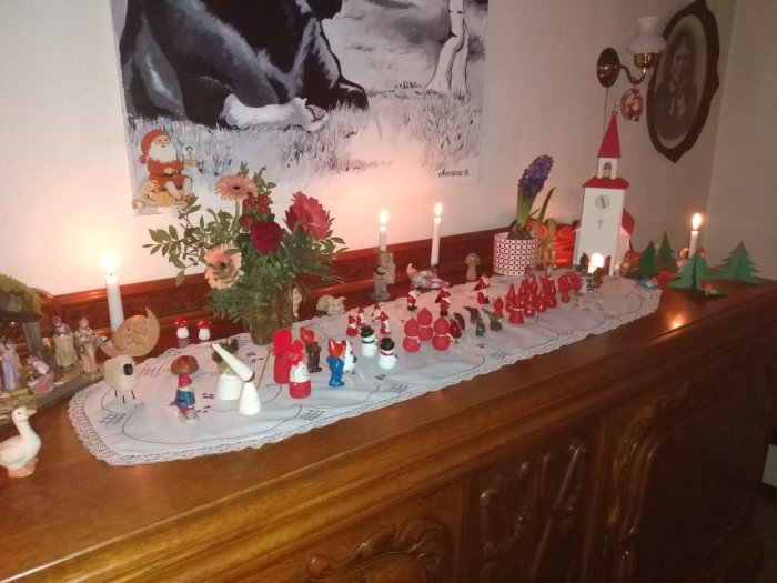 Juldekoration på en byrå med figurer, levande ljus, blommor och en tavla i bakgrunden.