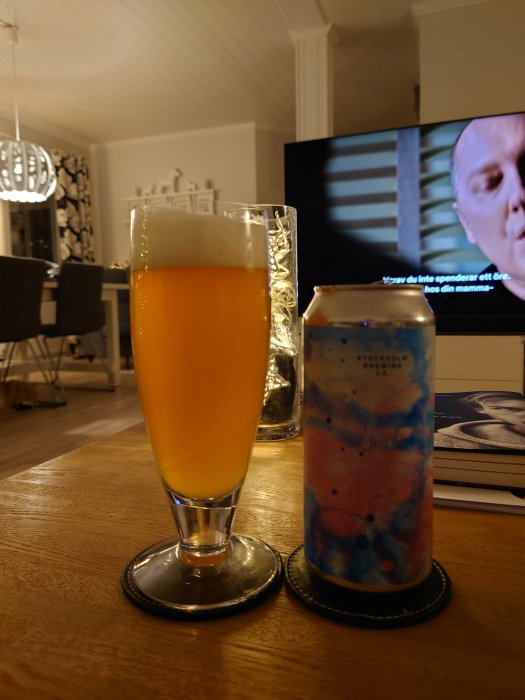 Ett glas öl och en ölburk på ett bord i ett vardagsrum, med en tv i bakgrunden.