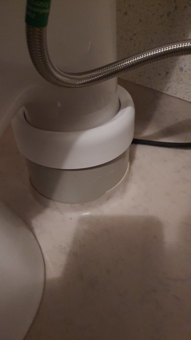 Vit gummimuff som förbinder WC-stol med avloppsrör, delvis täckt av plastkåpa.