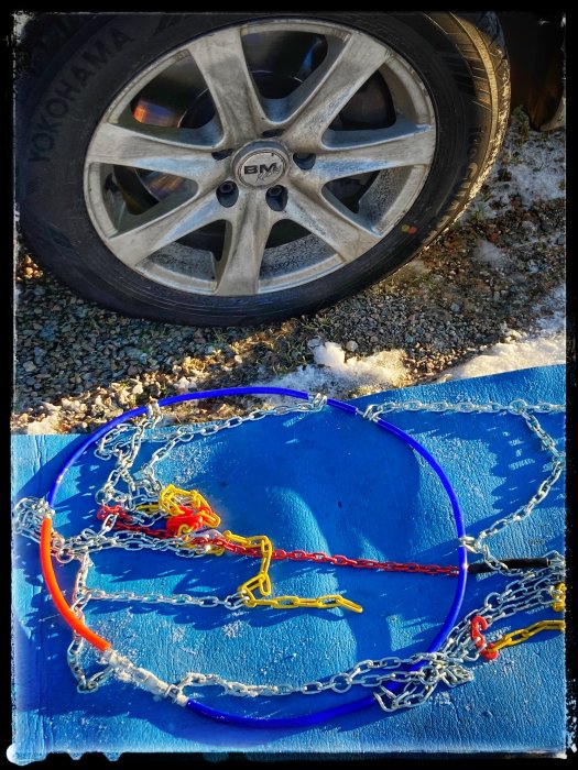 Bildäck med fälgar bredvid blå självspännande snökedjor på marken.