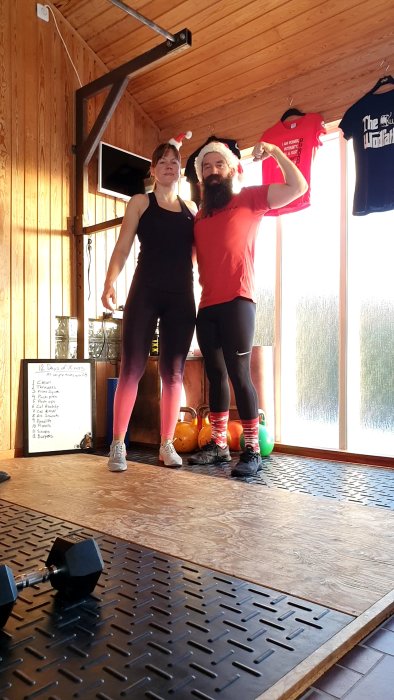 Två personer i träningskläder och julhattar poserar i ett hemmagym med kettlebells och hantel på golvet.