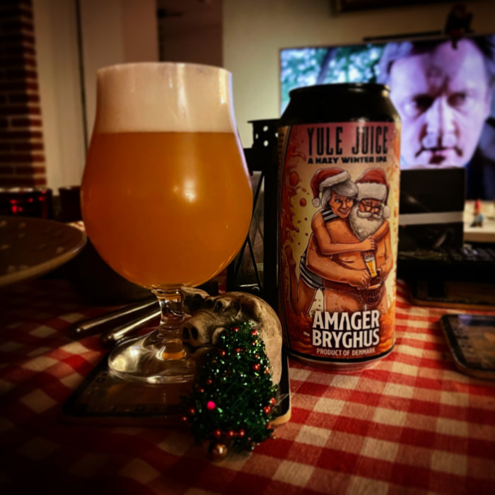 Ölglas fyllt med hazy IPA framför en burk märkt "Yule Juice", dekorerat gristryne och oskarp TV i bakgrunden.