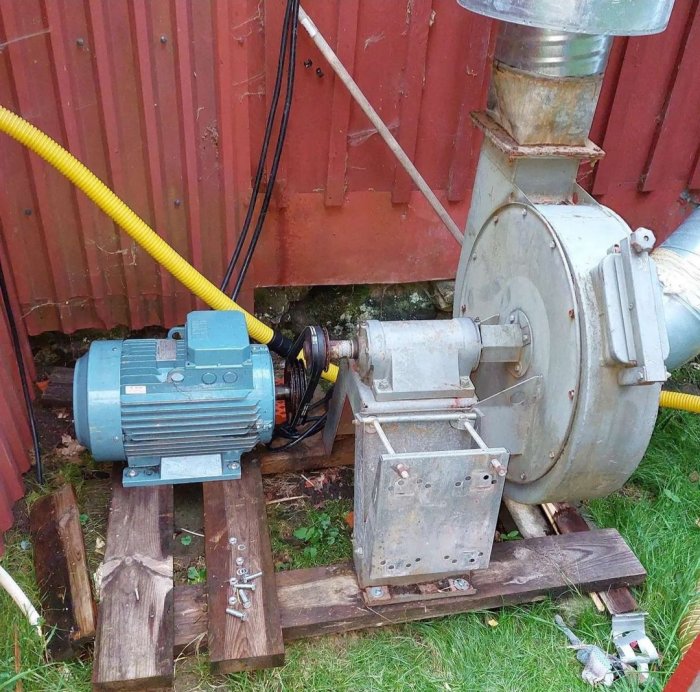 Tvåhastighets fläktmotor på träbalkar med ansluten gulkabel och avgasrör utomhus.