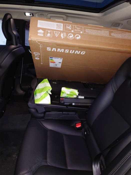 Samsung TV-kartong i ett bilbaksäte med säkerhetsbälte och gröna väskor.