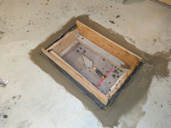Färskt gjutet betongfundament med träformar runtom och verktyg inuti på ett bygggolv.