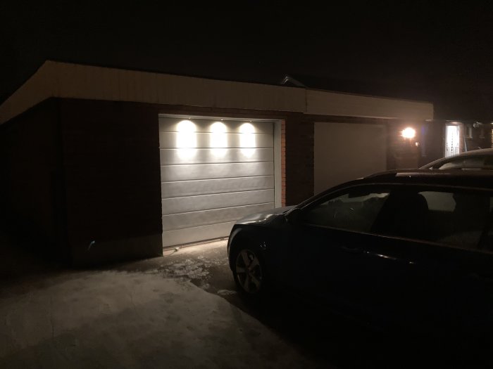 Nya spottar installerade ovanför garageporten i mörker, ger varmgult ljus på garage och bil.