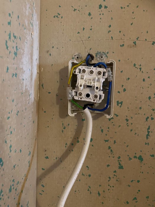 Oorganiserad elektrisk kabeldragning och öppen eluttag på en vägg med flagnande blå och gröna färger.
