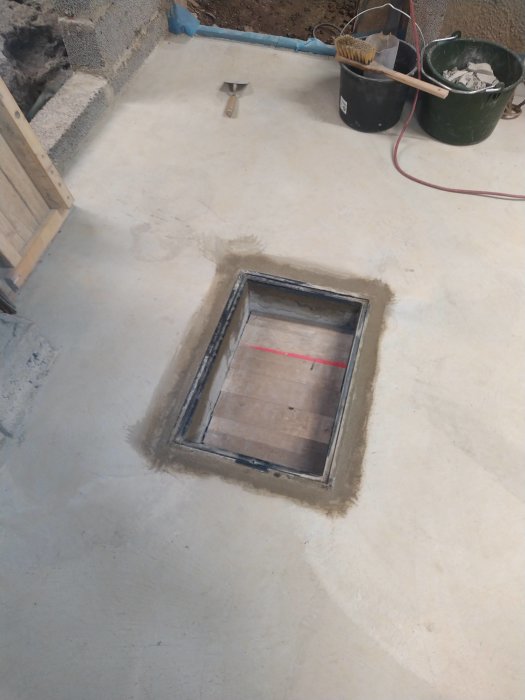 Nedsänkt pumpgropslucka i ett betonggolv med byggmaterial runtomkring.