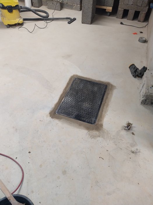 Integrerad pumpgropslucka i betonggolv med nätdesign, omgiven av byggmaterial och verktyg.