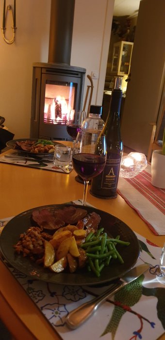 Middag framför öppen spis, med tallrik kött och grönsaker, vinflaska och glas vin.