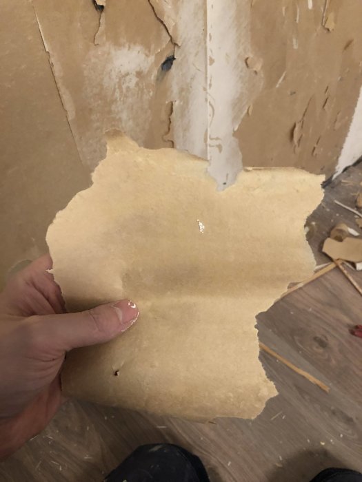 Hand håller en bit gammal gulaktig omslagsliknande papper med vägg i bakgrunden där bröstpanel har tagits bort.